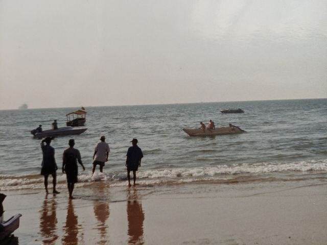 People wading at the water's edge at Calangute beach with small fishing boats offshore under a cloudy sky