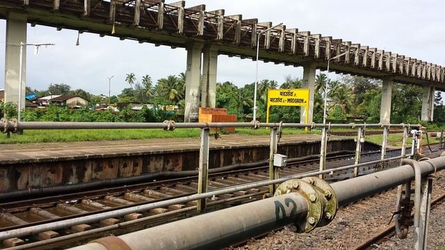 Railway tracks and a covered platform at Madgaon Junction station with a yellow station name board and palm trees