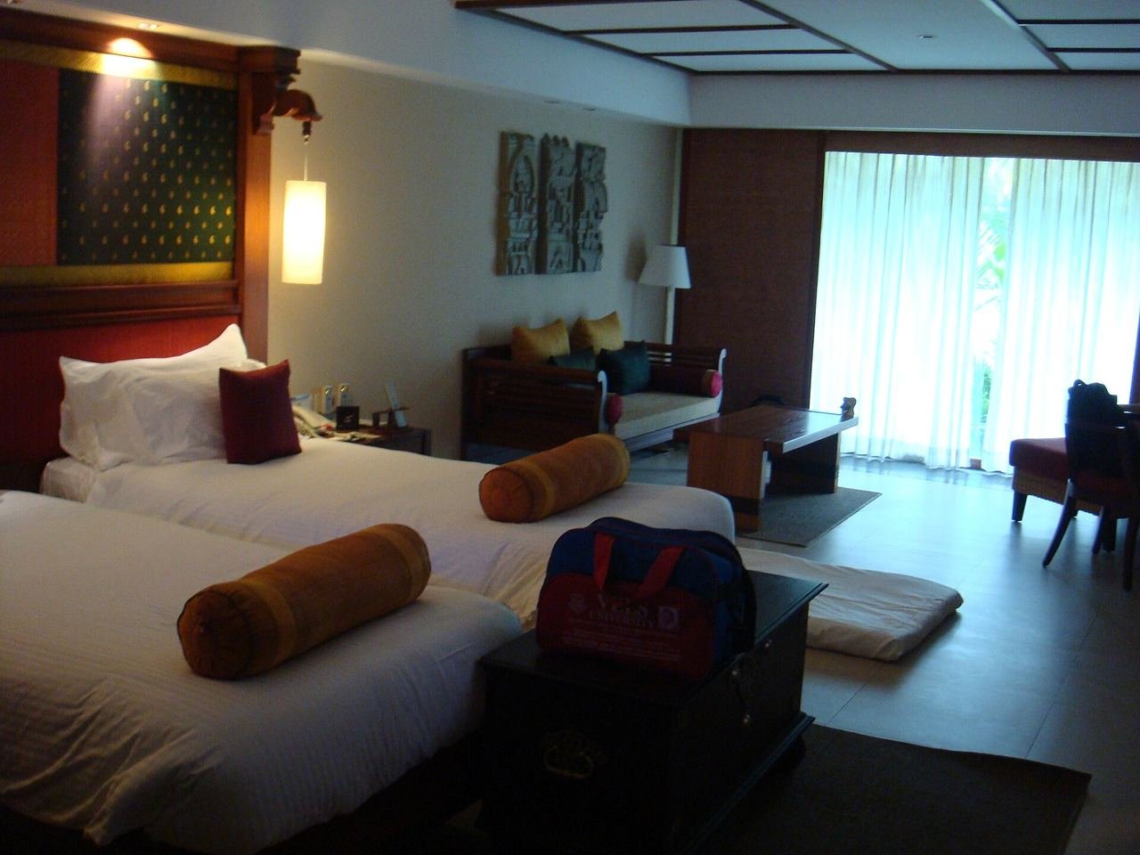 A hotel room with two made-up beds, bolster cushions, a sofa and a window with sheer curtains