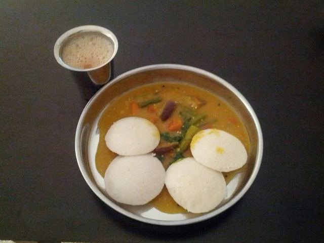 A steel plate of idli in sambar with a small steel tumbler of coffee on a dark table