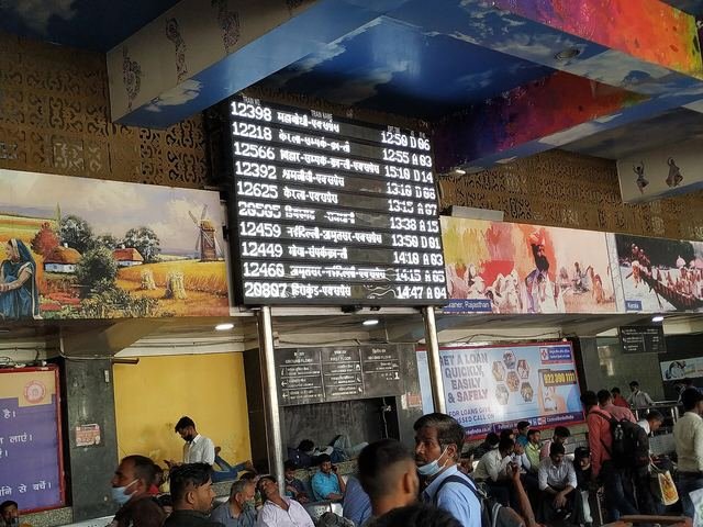 An electronic train departure board above waiting passengers in the hall of New Delhi railway station